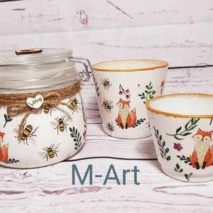 May include: Three white ceramic containers with a gold rim, decorated with a fox, bees, and floral designs. The containers are on a white wooden surface. The largest container is a glass jar with a lid and a twine bow. The lid has a small wooden heart with the word "Love" on it. The containers are labelled "M-Art".
