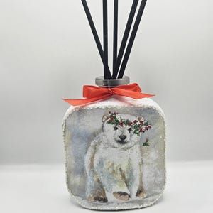 May include: A decorative reed diffuser featuring a polar bear illustration wearing a floral crown. The glass bottle is tied with a red ribbon and contains black reeds. The design has a winter theme.
