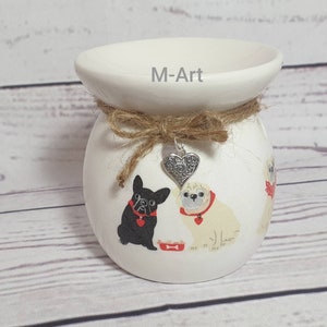 May include: White ceramic wax melt burner with a brown twine bow and a silver heart charm. The burner features a hand-painted design of three dogs, two pugs and a French bulldog, with red collars and a bone.