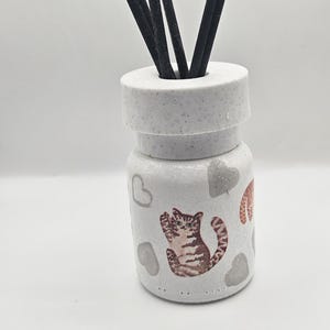 May include: White ceramic essential oil diffuser featuring a cat illustration and grey heart accents. The diffuser has a cylindrical shape with a round top and contains black reeds for fragrance dispersion. A decorative item for home fragrance.