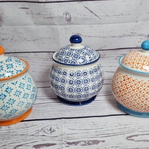 May include: Three ceramic canisters with lids. The canisters are white with blue and orange floral patterns. The lids are the same colour as the base of the canister.