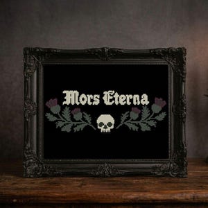 May include: Framed cross-stitch art with the words "Mors Eterna" above a skull and thistle design. The black frame sits on a wooden surface, with lit candles and a decorative vase in the background.