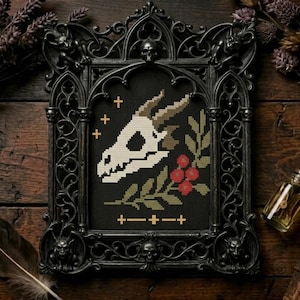 May include: A framed cross-stitch featuring a skull with horns, a sprig of berries, and gold crosses on a black background. The ornate black frame has gothic details, including skulls and gargoyles. The artwork is displayed on a wooden surface.
