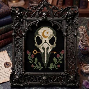 May include: A gothic-style framed cross-stitch featuring a skull with a crescent moon, keys, and floral accents. The black frame is adorned with gargoyles and skulls. The artwork is displayed on a dark surface with books and other gothic-themed items in the background.
