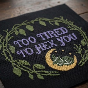 Puede incluir: Punto de cruz negro con el texto "TOO TIRED TO HEX YOU" en morado. Una rana verde descansa sobre una luna creciente amarilla, rodeada de estrellas. El diseño está enmarcado por una corona de hojas verdes.
