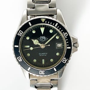 Puede incluir: Un reloj Hai plateado con una esfera negra y una correa plateada. El reloj tiene una ventana de fecha en la posición de las 3 en punto y un bisel giratorio. La esfera del reloj dice "Quartz 20 ATM."