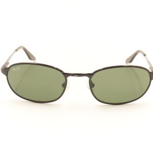 Pu&ograve; includere: Occhiali da sole neri con montatura ovale e lenti verdi. Gli occhiali hanno una montatura e aste in metallo sottile. Il logo Ray-Ban &egrave; visibile sulla lente superiore sinistra. Su sfondo bianco.