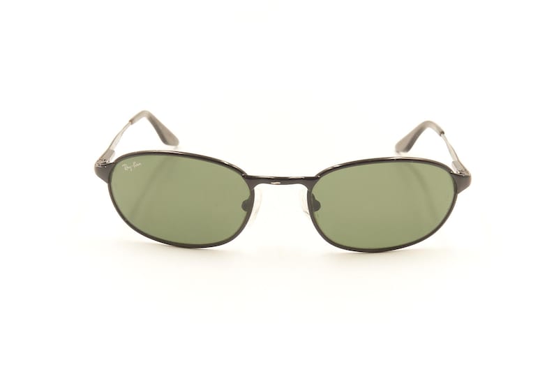 Pu&ograve; includere: Occhiali da sole neri con montatura ovale e lenti verdi. Gli occhiali hanno una montatura e aste in metallo sottile. Il logo Ray-Ban &egrave; visibile sulla lente superiore sinistra. Su sfondo bianco.