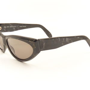 Pode incluir: &Oacute;culos de sol de tartaruga preta com arma&ccedil;&atilde;o curva e lentes marrons. Os &oacute;culos t&ecirc;m o texto "Bally Jan. U.S.A." na lateral.