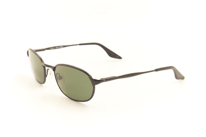 Pu&ograve; includere: Occhiali da sole ovali neri con lenti verdi. Le montature sono di colore metallico scuro e le aste sono sottili e dritte. Gli occhiali da sole hanno un design classico e sobrio, adatto all'uso quotidiano. Il testo "B&L RAY-BAN" &egrave; visibile sull'asta.