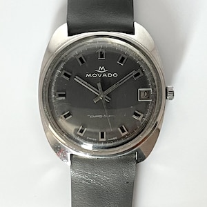 May include: A Movado watch with a dark gray face, silver case, and black leather band. The watch face features the Movado logo, rectangular hour markers, and a date window at the 3 o'clock position. The watch is attached to a gray leather strap.