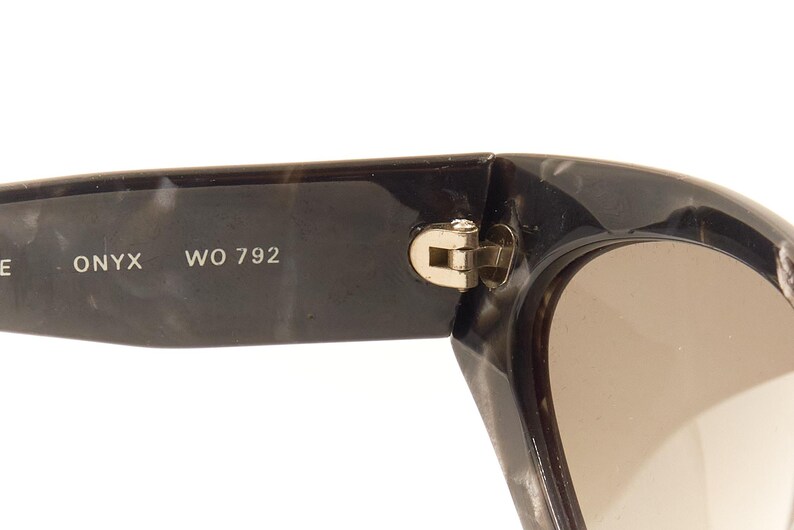 Pode incluir: &Oacute;culos de sol de tartaruga preto e cinza com dobradi&ccedil;a dourada. Os &oacute;culos t&ecirc;m lentes marrons e o texto "ONYX WO 792" est&aacute; impresso na arma&ccedil;&atilde;o.
