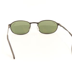 Pu&ograve; includere: Occhiali da sole ovali con montatura in metallo scuro e lenti verdi. Gli occhiali hanno un design classico. Le aste sono sottili e si estendono verso l'orecchio. Le lenti sono di colore verde chiaro. La montatura &egrave; grigio scuro.