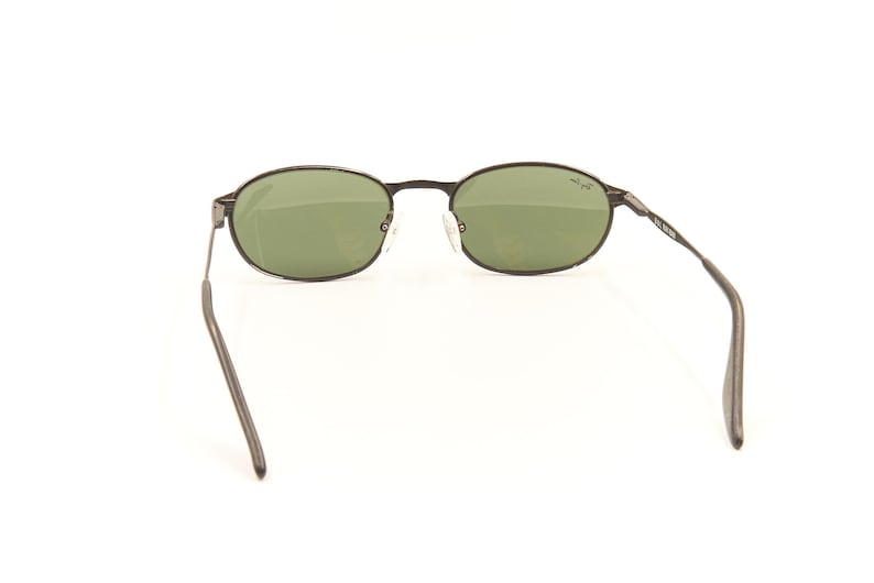 Pu&ograve; includere: Occhiali da sole ovali con montatura in metallo scuro e lenti verdi. Gli occhiali hanno un design classico. Le aste sono sottili e si estendono verso l'orecchio. Le lenti sono di colore verde chiaro. La montatura &egrave; grigio scuro.