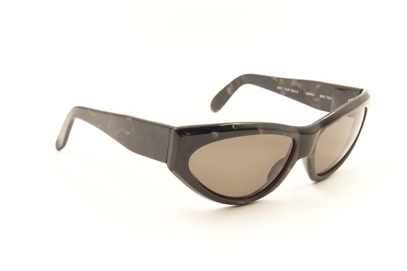 Pode incluir: &Oacute;culos de sol pretos de tartaruga com lentes marrons. Os &oacute;culos t&ecirc;m formato de olho de gato e uma arma&ccedil;&atilde;o fina.