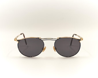 May include: A pair of round, black sunglasses with a gold metal frame. The sunglasses have a thin, wire-like frame and dark lenses.