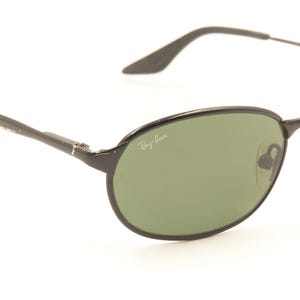 Pu&ograve; includere: Occhiali da sole ovali neri con lenti verdi. Il logo Ray-Ban &egrave; visibile sulla lente. Le aste sottili si estendono fino ai terminali. Gli occhiali da sole sono su uno sfondo bianco.