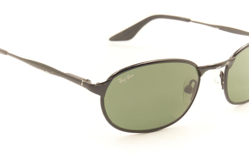 Pu&ograve; includere: Occhiali da sole ovali neri con lenti verdi. Il logo Ray-Ban &egrave; visibile sulla lente. Le aste sottili si estendono fino ai terminali. Gli occhiali da sole sono su uno sfondo bianco.