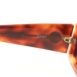 May include: Close-up of the tortoiseshell frame of a pair of sunglasses with a gold hinge. The sunglasses have a brown tortoiseshell frame and amber lenses. The frame has the number W2527 printed on it.