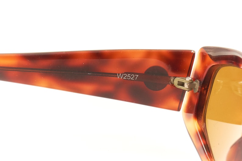 May include: Close-up of the tortoiseshell frame of a pair of sunglasses with a gold hinge. The sunglasses have a brown tortoiseshell frame and amber lenses. The frame has the number W2527 printed on it.