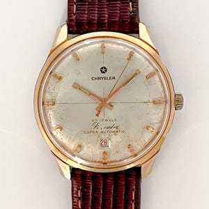 Op de afbeelding: Een goudkleurige Chrysler-horloge met een witte wijzerplaat en een bruin leren bandje. De wijzerplaat van het horloge heeft het Chrysler-logo, de tekst "25 Jewels", "Kr. cal. 17", "Super-Automatic" en "28".