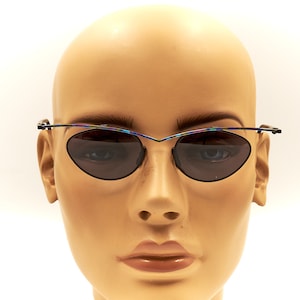May include: A pair of oval-shaped sunglasses with a thin, iridescent metal frame. The sunglasses have dark gray lenses and are worn by a mannequin.