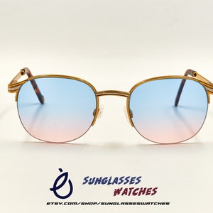 Op de afbeelding: Gouden zonnebril met een blauwe tot roze verlooplens. De zonnebril heeft een dun, gouden metalen montuur.