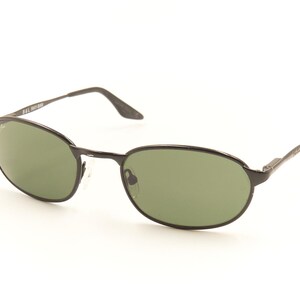 Pu&ograve; includere: Occhiali da sole ovali neri con lenti verdi. Gli occhiali hanno una sottile montatura e aste in metallo nero. Il logo Ray-Ban &egrave; visibile sulla lente e sull'asta sinistra. Gli occhiali sono su sfondo bianco.