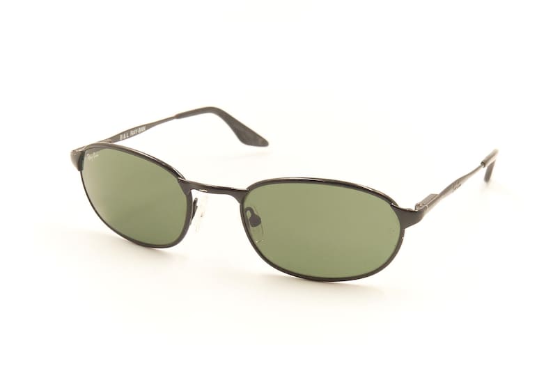 Pu&ograve; includere: Occhiali da sole ovali neri con lenti verdi. Gli occhiali hanno una sottile montatura e aste in metallo nero. Il logo Ray-Ban &egrave; visibile sulla lente e sull'asta sinistra. Gli occhiali sono su sfondo bianco.