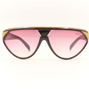 Może przedstawiać: Okulary przeciwsłoneczne w stylu vintage z czarną oprawką i różowymi soczewkami gradientowymi. Górna część oprawki ma brązowy wzór słojów drewna ze złotymi akcentami. Okulary przeciwsłoneczne mają unikalny, duży kształt.