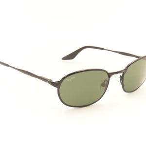 Pu&ograve; includere: Occhiali da sole ovali neri con lenti verdi. Gli occhiali hanno una montatura e aste sottili in metallo nero. Il logo Ray-Ban &egrave; visibile sulla lente. Gli occhiali sono su sfondo bianco.
