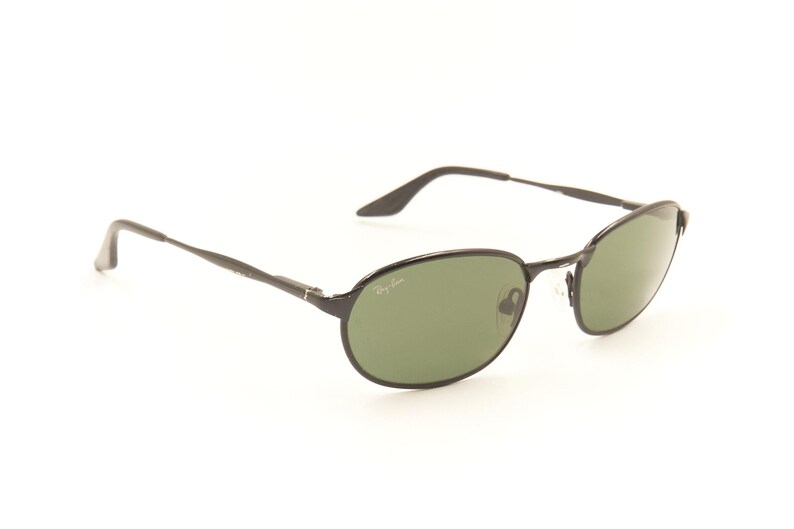 Pu&ograve; includere: Occhiali da sole ovali neri con lenti verdi. Gli occhiali hanno una montatura e aste sottili in metallo nero. Il logo Ray-Ban &egrave; visibile sulla lente. Gli occhiali sono su sfondo bianco.
