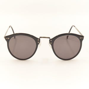 May include: Black round sunglasses with gray lenses. The frames are black with silver accents at the hinges. The temples are thin and silver. The sunglasses are set against a white background.