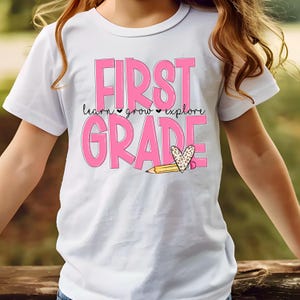 May include: White t-shirt with the words "FIRST GRADE" in pink block letters. Below the word "FIRST" are the words "learn, grow, explore" in a smaller, black font. A pencil and heart graphic are below the word "GRADE".