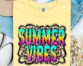 May include: A light yellow t-shirt with the words "SUMMER VIBES" in a colorful, gradient font. The text is outlined in black and set against a leopard print background. The shirt is folded on a light surface.
