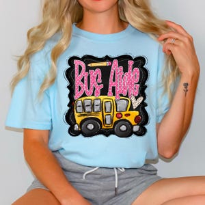 May include: Light blue t-shirt featuring a graphic of a yellow school bus and the words "Bus Aide" in pink bubble letters. The design includes a pencil and a heart. The shirt is made of a soft material.