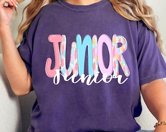 May include: A purple t-shirt with the word "JUNIOR" in large, colorful letters across the front. The word "junior" is written in a smaller, cursive font below the main word. The letters are pink, blue, and yellow.