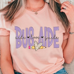 Op de afbeelding: Lichtroze T-shirt met de woorden "BUS AIDE" in een paarse, omrande letter. De woorden "aide love guide" zijn in een zwart script geschreven. Een potlood en een hart met een luipaardprint zijn ook afgebeeld.