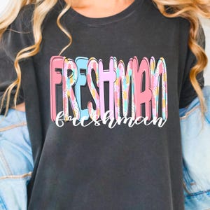 Könnte beinhalten: Ein dunkelgraues T-Shirt mit dem Wort "FRESHMAN" in großen, bunten Buchstaben. Unter dem Wort "FRESHMAN" steht das Wort "freshman" in einer kursiven Schriftart. Die Buchstaben sind rosa, blau und gelb.