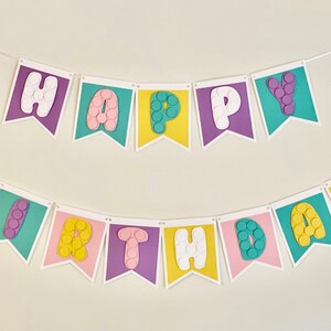 Pode incluir: Uma faixa de feliz anivers&aacute;rio colorida com bandeiras roxas, amarelas, cor-de-rosa e azul-turquesa. Cada letra &eacute; feita de papel branco com um design texturizado.