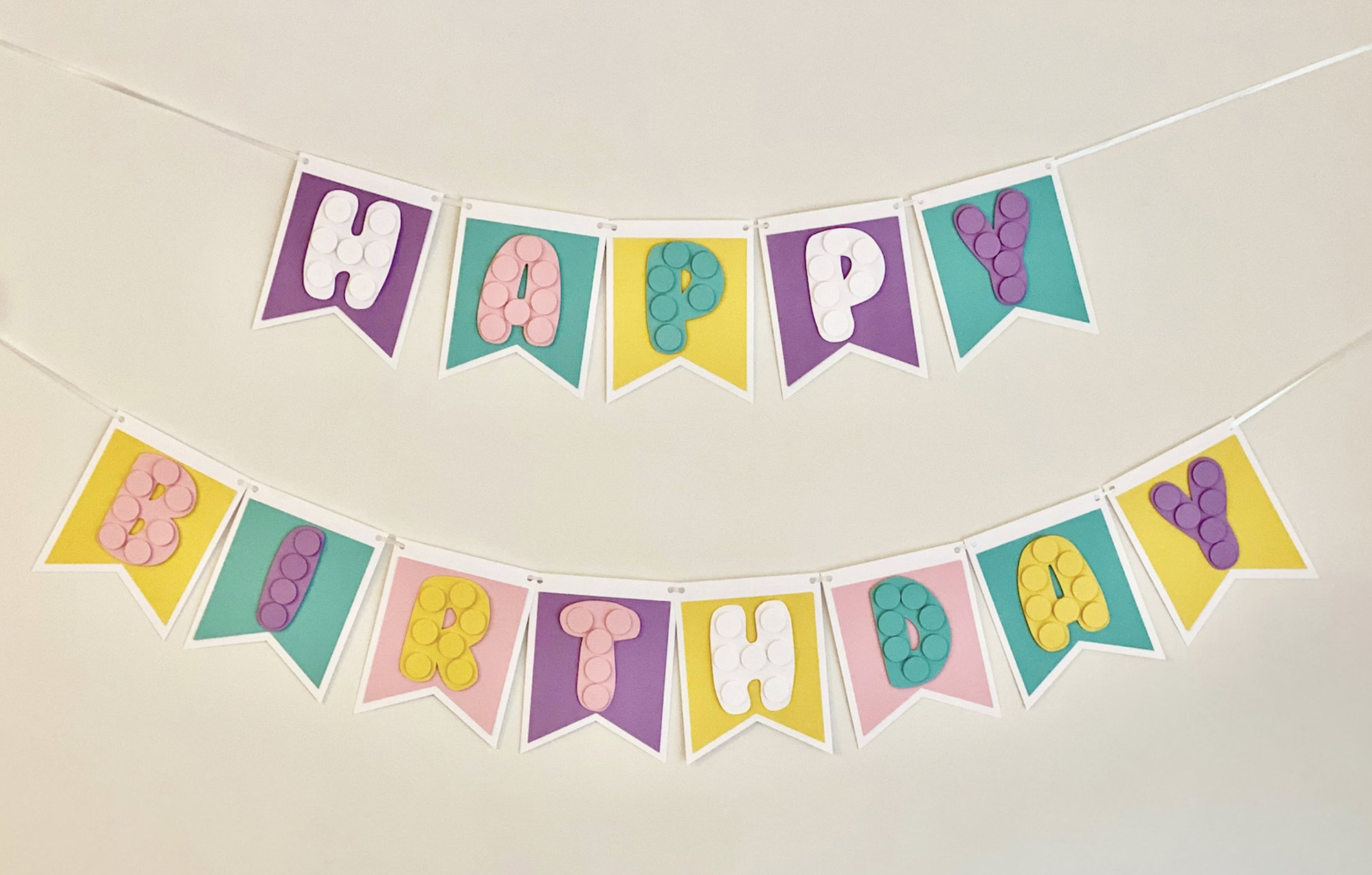 Pode incluir: Uma faixa de feliz anivers&aacute;rio colorida com bandeiras roxas, amarelas, cor-de-rosa e azul-turquesa. Cada letra &eacute; feita de papel branco com um design texturizado.