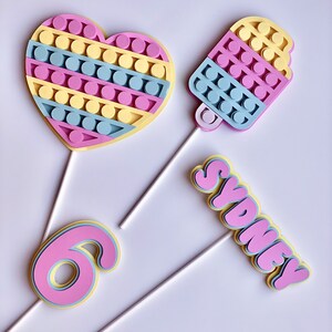 Pode incluir: Tr&ecirc;s toppers para bolo com um cora&ccedil;&atilde;o, um cone de sorvete e o nome "Sydney" em fonte de bolha. Todos os tr&ecirc;s toppers s&atilde;o feitos de espuma rosa, amarela e azul e t&ecirc;m um palito branco preso na parte de tr&aacute;s.