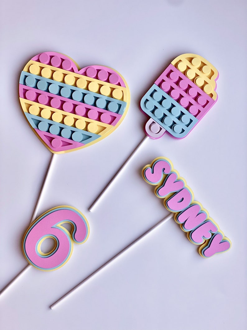 Pode incluir: Tr&ecirc;s toppers para bolo com um cora&ccedil;&atilde;o, um cone de sorvete e o nome "Sydney" em fonte de bolha. Todos os tr&ecirc;s toppers s&atilde;o feitos de espuma rosa, amarela e azul e t&ecirc;m um palito branco preso na parte de tr&aacute;s.