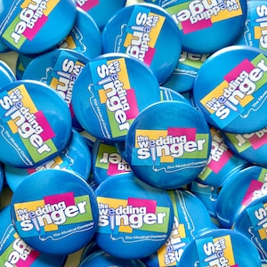May include: A pile of blue round buttons with the words "The Wedding Singer" and "The Musical Comedy" in colourful text.