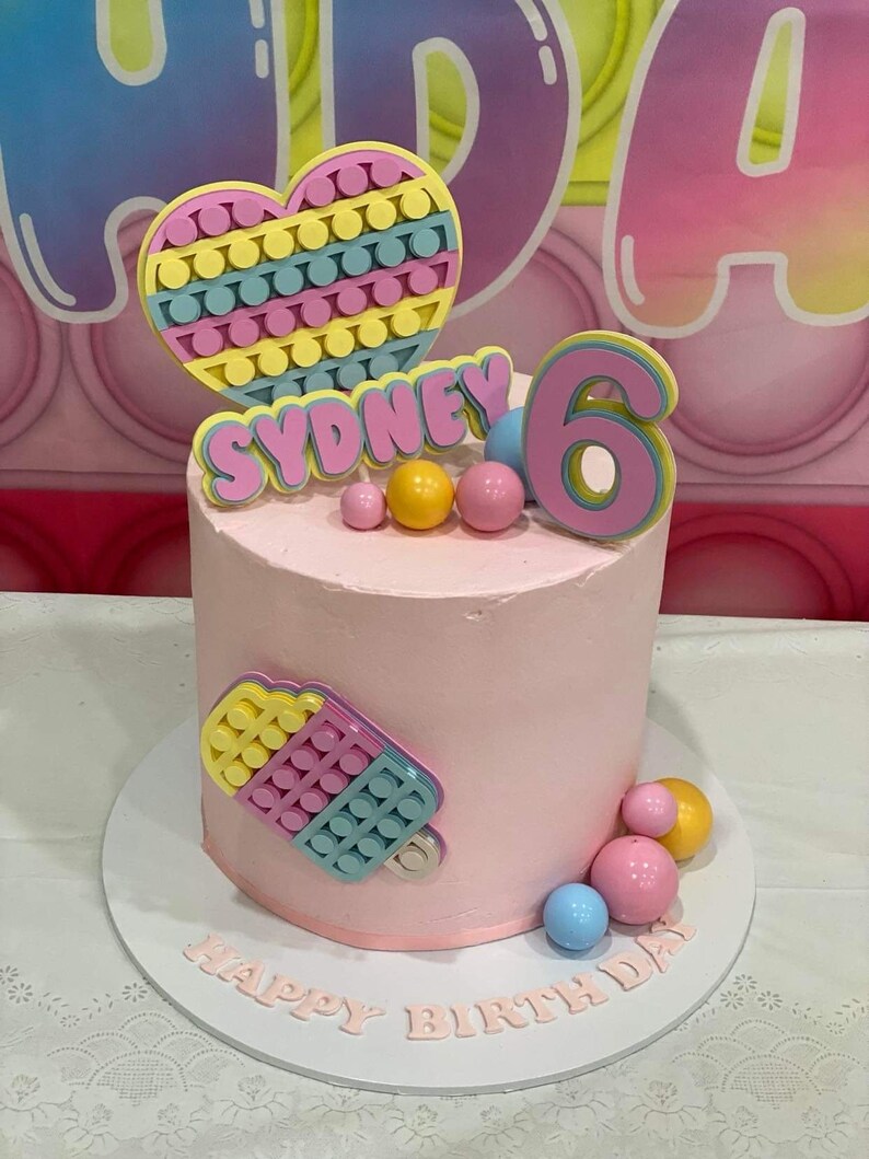 Pode incluir: Um bolo rosa decorado com um brinquedo pop-it em forma de cora&ccedil;&atilde;o, um n&uacute;mero 6 rosa e o nome "Sydney". O bolo &eacute; cercado por brinquedos pop-it coloridos e as palavras "Feliz anivers&aacute;rio" est&atilde;o escritas no suporte do bolo.