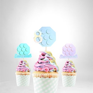 Pode incluir: Tr&ecirc;s cupcakes com cobertura rosa pastel, azul e amarela. Cada cupcake tem um topo decorativo com o n&uacute;mero 8 e uma forma hexagonal com c&iacute;rculos. Os topos s&atilde;o em azul, rosa e azul claro. Os cupcakes est&atilde;o em forminhas xadrez verdes e brancas. Os topos dizem "Savannah" em dois deles.