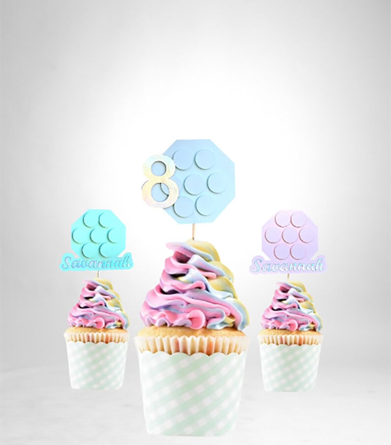 Pode incluir: Tr&ecirc;s cupcakes com cobertura rosa pastel, azul e amarela. Cada cupcake tem um topo decorativo com o n&uacute;mero 8 e uma forma hexagonal com c&iacute;rculos. Os topos s&atilde;o em azul, rosa e azul claro. Os cupcakes est&atilde;o em forminhas xadrez verdes e brancas. Os topos dizem "Savannah" em dois deles.