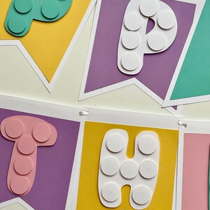 Pode incluir: Uma faixa colorida com a palavra "Happy" escrita em letras brancas com c&iacute;rculos brancos em um fundo roxo, amarelo e verde.
