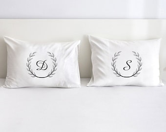 May include: Two white pillows featuring black monogram letters 'D' and 'S' within a laurel wreath design. The pillows are placed on a white bed, offering a clean, minimalist look.