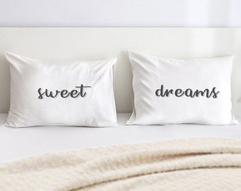 May include: Two white pillows with the words "sweet" and "dreams" printed in dark grey script. The pillows are on a white bed with a beige blanket draped over the edge.